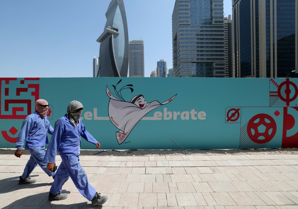 Des travailleurs passent devant une palissade de chantier où l'on voit la mascotte La'eeb de la Coupe du monde de football au Qatar, le 13 octobre 2022 à Doha